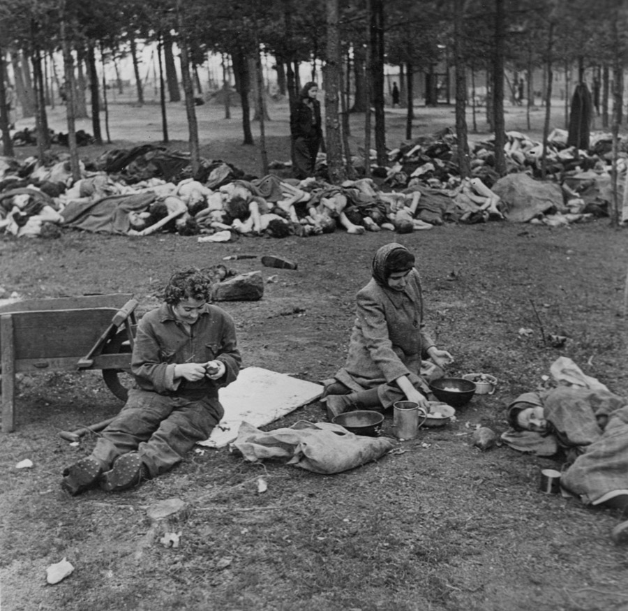 Women peeling potatoes; in the background are piles of dead bodies